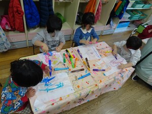 3歳児4名が1つの机の周りに座り、一人1枚ずつビニールに様々な色の油性ペンで絵を描いています